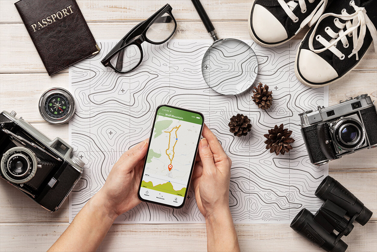 Frau hält Handy, auf dem eine Routen-App geöffnet ist, um sie herum liegen Reiseutensilien.